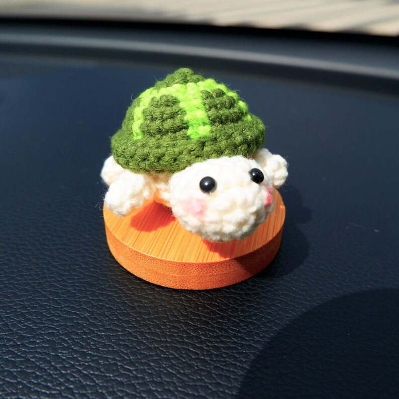 May include: A handmade crochet turtle figurine. The turtle has a green and light green shell, a white head and limbs, and black eyes. It sits on a small, round, orange wooden base. The turtle's cheeks are lightly blushed pink.
