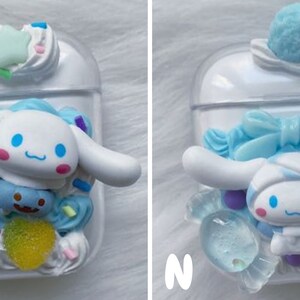 May include: Two white Airpod cases decorated with blue and white 3D characters, candy, and sprinkles. One case features a blue bow and the other features a safety pin.