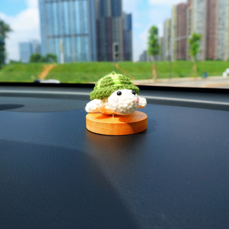 May include: A crocheted turtle figurine with a green shell and white body sits on a small wooden base. The turtle has black eyes and pink accents. The figurine is displayed on a dark dashboard with a blurred cityscape in the background.