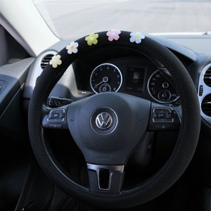 Puede incluir: Funda de volante negra con detalles de flores bordadas de colores. La funda está diseñada para adaptarse a un volante de coche estándar. Las flores son en tonos amarillo, rosa, azul y blanco. El salpicadero y los controles del coche son visibles.