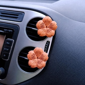 May include: Two wooden, flower-shaped car air vent diffusers. The diffusers are a warm brown color and are placed on the air vents of a car's dashboard. The dashboard is silver and black.