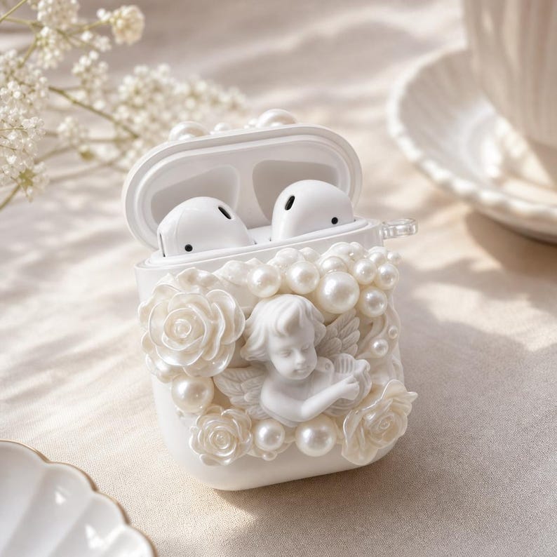 May include: White AirPods case decorated with pearls, roses, and a cherub. The case is open, revealing two white earbuds. The design is ornate and elegant, with a focus on the pearl and floral details.