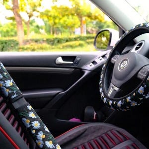 May include: A black steering wheel cover and a matching seat belt shoulder pad cover, both featuring a repeating pattern of white daisy flowers with yellow centers and green stems against a dark background, installed inside a vehicle.