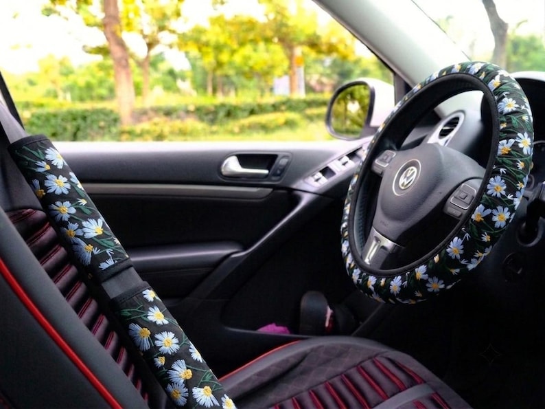 May include: A black steering wheel cover and a matching seat belt shoulder pad cover, both featuring a repeating pattern of white daisy flowers with yellow centers and green stems against a dark background, installed inside a vehicle.