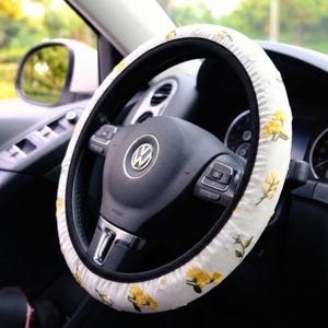 Può includere: Coprivolante bianco con ricami floreali gialli. La copertura è su un volante nero con il logo Volkswagen argentato. L'interno dell'auto comprende uno specchietto laterale e un cruscotto.