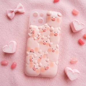 Puede incluir: Una funda de teléfono rosa con un diseño 3D de varios cerdos de dibujos animados. Los cerdos tienen mejillas rosadas y pequeños ojos negros. También se ven un lazo rosa y malvaviscos en forma de corazón.