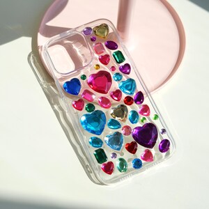 May include: Clear phone case adorned with colorful, jewel-like embellishments. The case features heart, square, and oval-shaped gems in shades of blue, pink, green, purple, and gold. The design is eye-catching and adds a touch of sparkle.