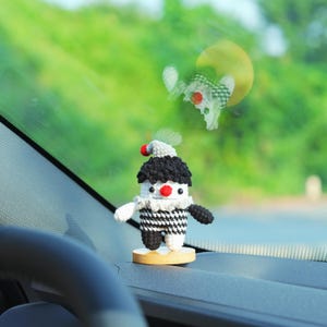 May include: A handmade, crocheted clown doll with a black and white checkered body, black hair, and a red nose. The doll is mounted on a small wooden base and is designed as a car accessory or decorative item.