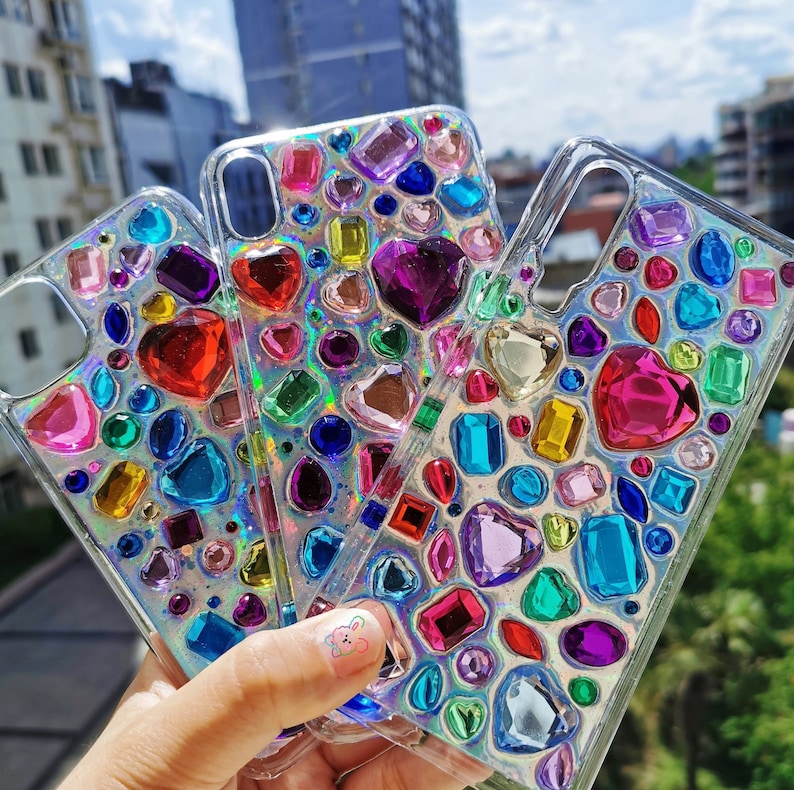 May include: Three clear phone cases adorned with colorful, iridescent gemstones in various shapes. The cases feature heart, square, and oval-shaped gems in shades of red, pink, purple, blue, and green. The cases are held up against a bright, sunny background.