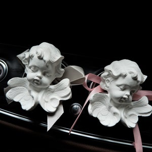 May include: Two white cherub figurines with wings, each with a pink ribbon tied around their necks. The cherubs are placed on a black dashboard of a car.