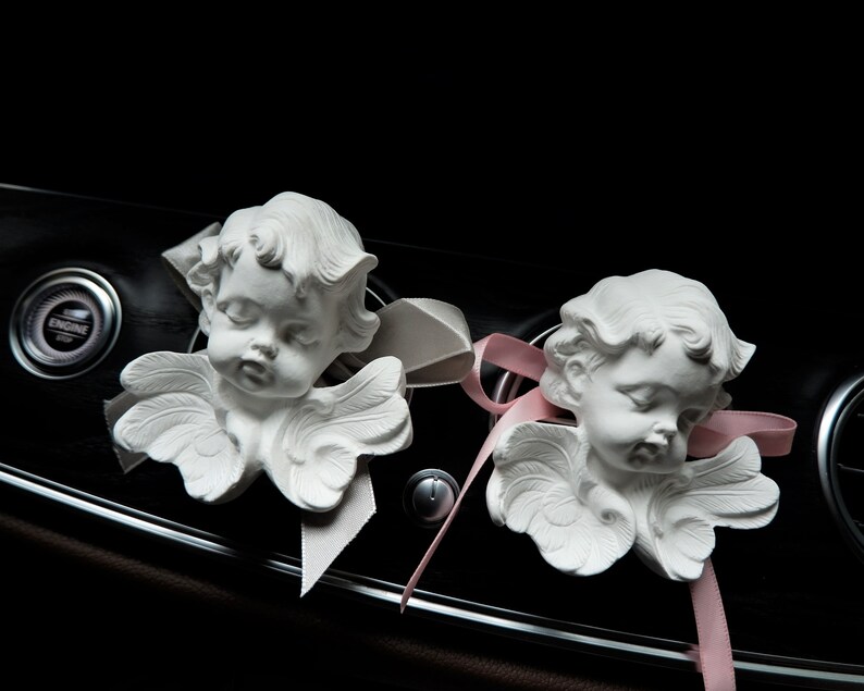 May include: Two white cherub figurines with wings, each with a pink ribbon tied around their necks. The cherubs are placed on a black dashboard of a car.