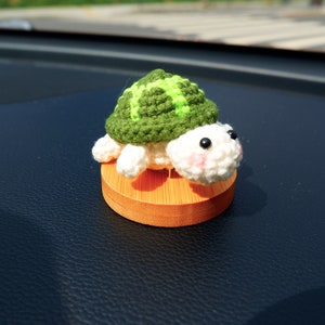 May include: A handmade crochet turtle figurine. The turtle has a green shell with a lighter green pattern, white head and limbs, and black eyes. It sits on a small, round, wooden base. The turtle's face has pink blush.