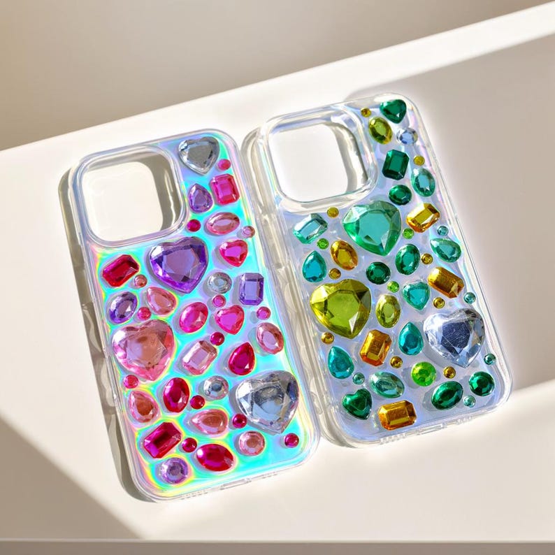 May include: Two clear phone cases adorned with colorful, iridescent gemstones. The cases feature heart, oval, and rectangular gems in shades of pink, purple, green, and yellow. The cases are displayed on a white surface.