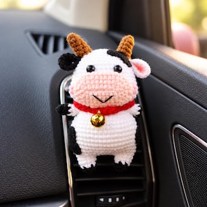 Può includere: Una statuetta di mucca all'uncinetto con corpo bianco, macchie nere e corna marroni. La mucca ha un muso rosa, occhi neri e un collare rosso con un piccolo campanello dorato lucido. La figura è fissata a una bocchetta di ventilazione dell'auto.