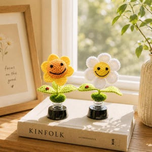 May include: Two crocheted flower ornaments with smiling faces. One is yellow, the other white, both with green leaves and a black base. They sit on a book that says "KINFOLK". A framed print with the words "focus on the good" is in the background.