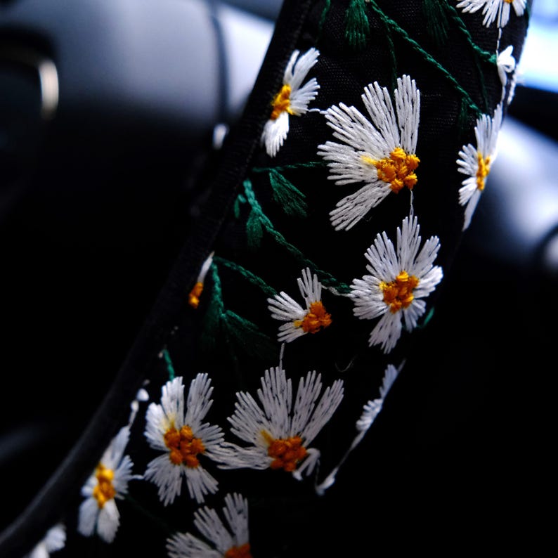 May include: A black steering wheel cover featuring an embroidered pattern of white daisy flowers with yellow centers and green stems. The floral design is stitched directly onto the dark fabric, creating a textured surface across the cover.