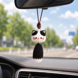 Op de afbeelding: Een handgemaakte auto-hanger met een zwart-wit kattenontwerp. De kat heeft een witte kop met zwarte oren en vlekken, roze wangen en kleine snorharen. Het lichaam is zwart met een witte onderkant. Het hangt aan een bruin leren koord.
