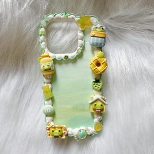 May include: A decorative phone case with a light green and white border. The case is adorned with various 3D embellishments, including a frog in a jar, a pineapple, a strawberry, and a house. The case has a light green background.