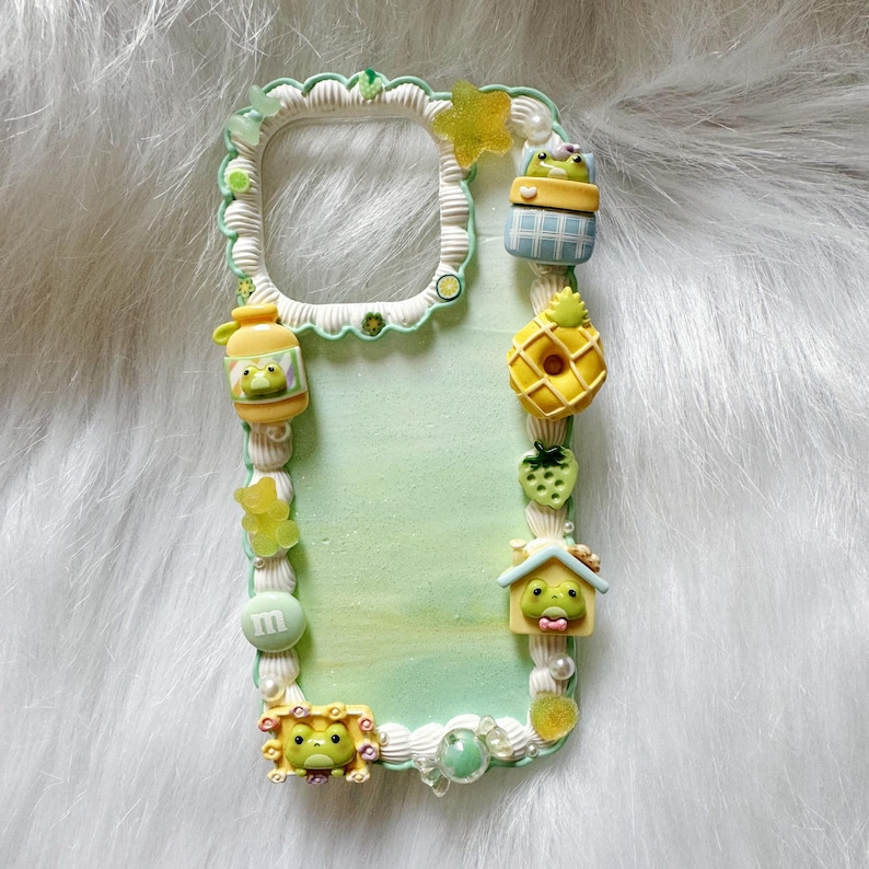 May include: A decorative phone case with a light green and white border. The case is adorned with various 3D embellishments, including a frog in a jar, a pineapple, a strawberry, and a house. The case has a light green background.