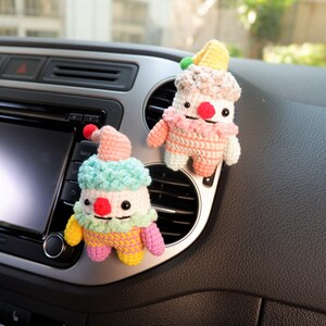 May include: Two crocheted clown dolls made with colourful yarn and smiling faces hanging from the air vent of a car.