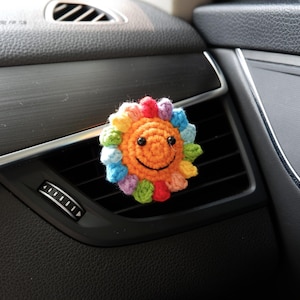 May include: A colourful, crocheted sun-shaped car air vent clip. The sun has an orange centre with a smiling face and rainbow-coloured rays. The clip is attached to a black car vent, adding a cheerful touch to the vehicle's interior.