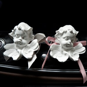 May include: Two white porcelain cherub figurines with wings, each with a pink ribbon tied around their necks. The cherubs are placed on a black dashboard with a pink ribbon extending to the right.
