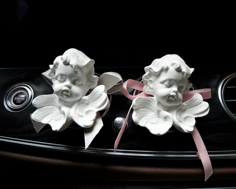 May include: Two white porcelain cherub figurines with wings, each with a pink ribbon tied around their necks. The cherubs are placed on a black dashboard with a pink ribbon extending to the right.