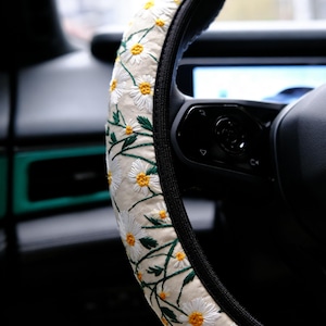 May include: A steering wheel cover featuring a cream-colored fabric base with an embroidered pattern of white daisies with yellow centers and green stems. The cover has a black trim along the inner edge and is fitted onto a black steering wheel.