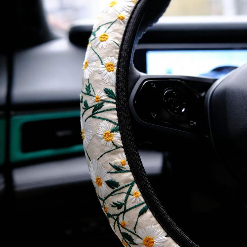 May include: A steering wheel cover featuring a cream-colored fabric base with an embroidered pattern of white daisies with yellow centers and green stems. The cover has a black trim along the inner edge and is fitted onto a black steering wheel.