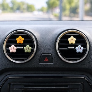 Op de afbeelding: Vijf kleine, gehaakte stervormige ornamenten bevestigd aan de horizontale lamellen van twee ronde ventilatieroosters in een auto. De sterren zijn oranje, lichtroze, lichtgroen, lichtgeel en lichtblauw, elk voorzien van geborduurde ogen en een glimlach.