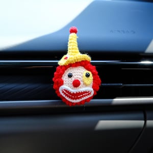 Puede incluir: Una cara de payaso hecha a mano con ganchillo, con una combinación de colores rojo, amarillo y blanco. El payaso tiene un afro rojo, un sombrero amarillo y una cara blanca con ojos negros, una nariz roja y una sonrisa roja y blanca. Accesorio de ventilación.