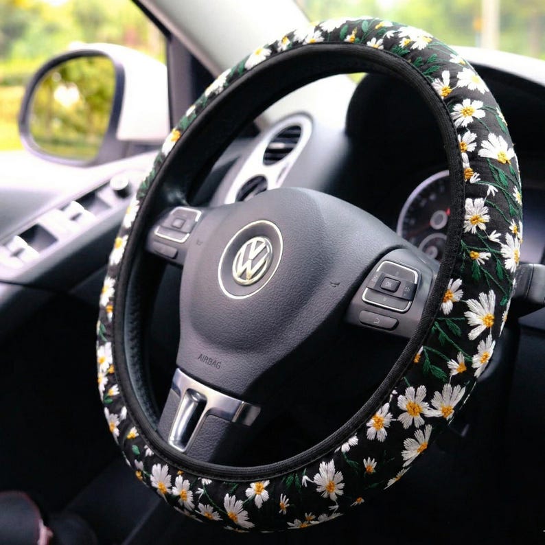 May include: A black steering wheel cover featuring an embroidered pattern of white daisy flowers with yellow centers and green stems. The cover is fitted onto a black Volkswagen steering wheel with a central silver logo and integrated control buttons.