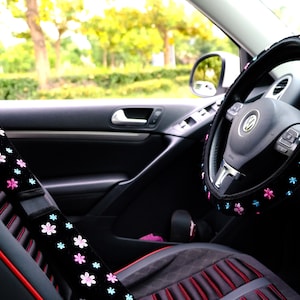 May include: A black car interior with a black steering wheel cover with pink and blue flowers. The steering wheel cover is made of soft fabric and has a comfortable grip. The car seat is black with red stitching and a black seatbelt cover with pink and blue flowers.