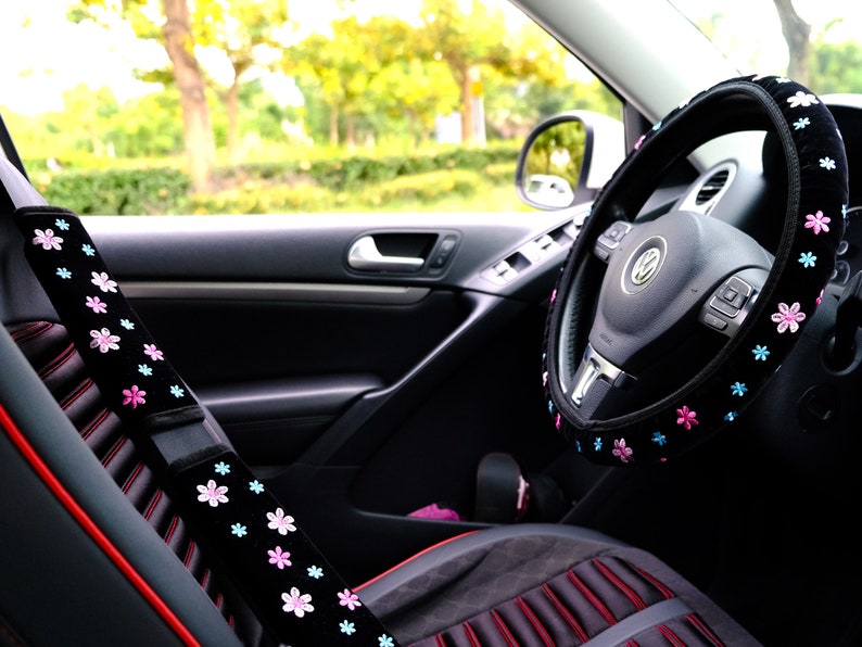 May include: A black car interior with a black steering wheel cover with pink and blue flowers. The steering wheel cover is made of soft fabric and has a comfortable grip. The car seat is black with red stitching and a black seatbelt cover with pink and blue flowers.
