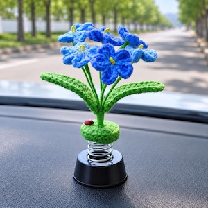 May include: A crocheted blue flower ornament with green leaves and a small red ladybug accent. The flower is attached to a green crocheted base, which sits on a silver metal spring connected to a smooth black circular stand for dashboard display.