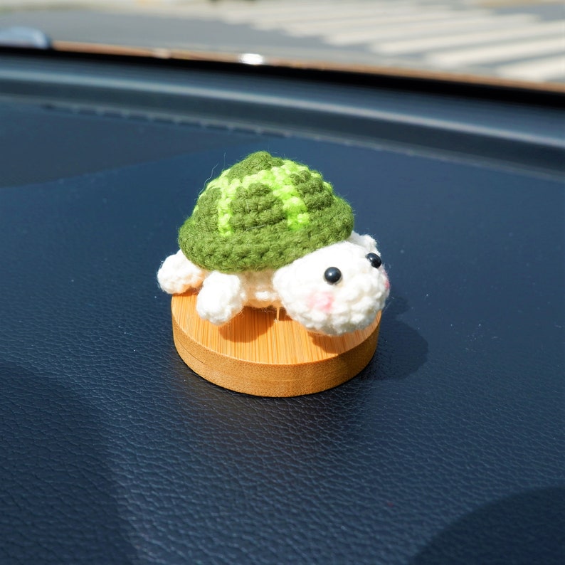May include: A handmade crochet turtle figurine with a green shell and white body. The turtle has black eyes and pink cheeks, and sits on a small, round wooden base. The turtle is a cute, decorative item.