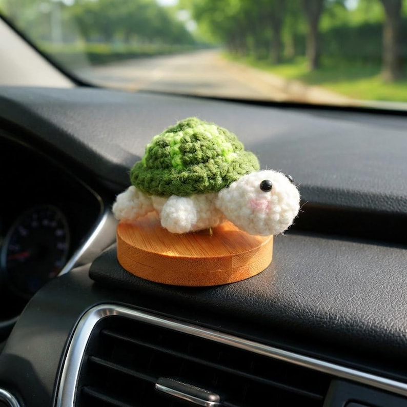 May include: A handmade crochet turtle figurine with a green and white shell, white body, and black eyes. The turtle is mounted on a small, round wooden base. The figurine is displayed on a car dashboard.