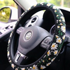 May include: A black steering wheel cover featuring an embroidered pattern of white daisies with yellow centers and green stems. The cover is fitted onto a black Volkswagen steering wheel with a central silver logo and integrated control buttons.