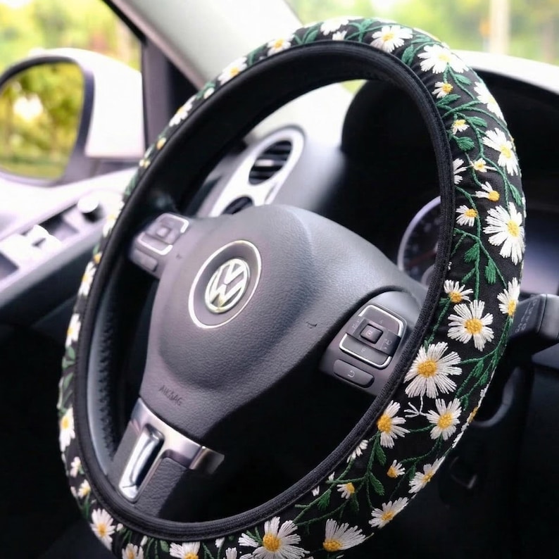 May include: A black steering wheel cover featuring an embroidered pattern of white daisies with yellow centers and green stems. The cover is fitted onto a black Volkswagen steering wheel with a central silver logo and integrated control buttons.