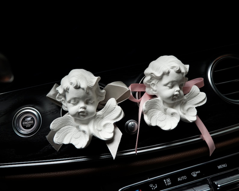 May include: Two white ceramic angel figurines with wings, placed on a black car dashboard with a pink ribbon. The angel figurines have closed eyes and are facing forward.
