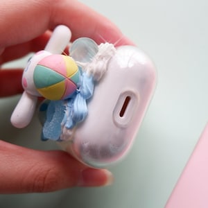 May include: A white Airpods case with a 3D design of a blue and white ice cream cone with a pink and yellow beach ball on top. The case has a charging port on the bottom.