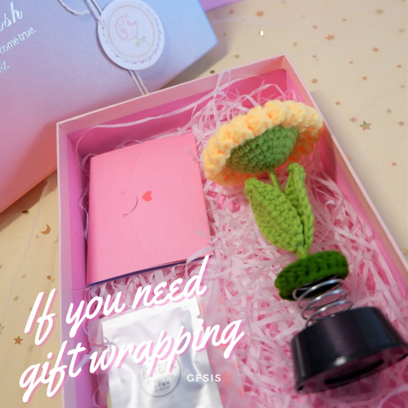 May include: A pink gift box with white tissue paper, a pink card with a heart, and a crocheted yellow sunflower on a spring base. The text "If you need gift wrapping" is visible on the box.