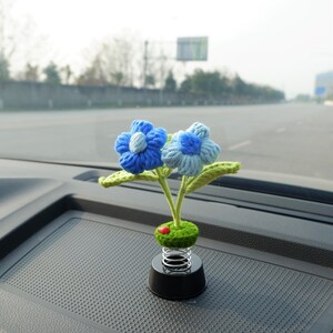 May include: A blue and white crocheted flower car air freshener with green leaves and a spring base. The air freshener is sitting on a black dashboard.