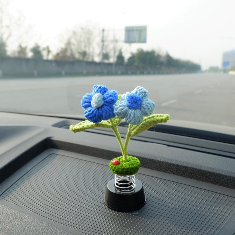 May include: A blue and white crocheted flower car air freshener with green leaves and a spring base. The air freshener is sitting on a black dashboard.