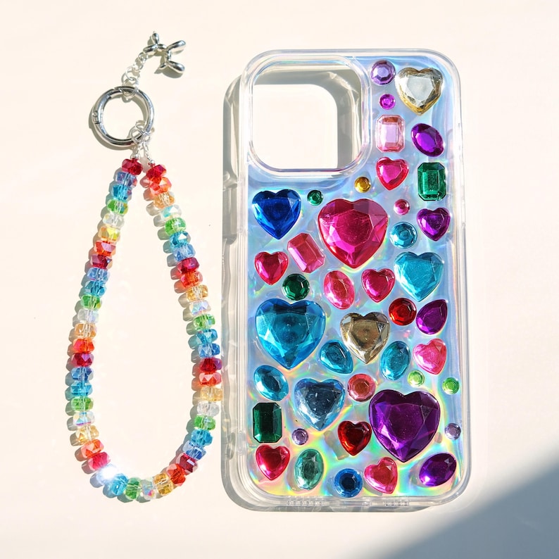 May include: Clear phone case adorned with colorful heart-shaped and rectangular gems. A rainbow beaded wrist strap with a silver-tone clasp is attached. The case features various jewel tones, including pink, blue, and green.