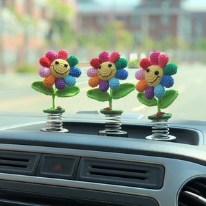 May include: Three colorful crocheted flower car dashboard decorations with smiling faces. Each flower is attached to a spring.