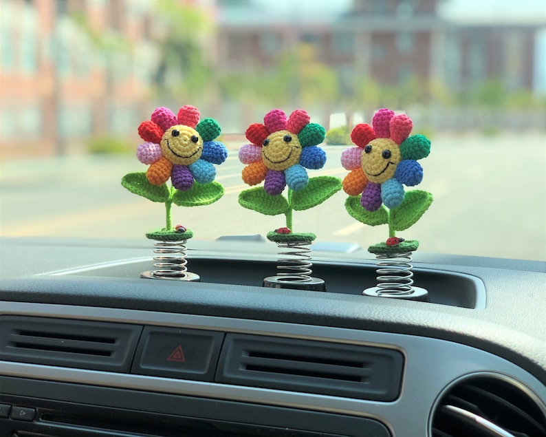 May include: Three colorful crocheted flower car dashboard decorations with smiling faces. Each flower is attached to a spring.