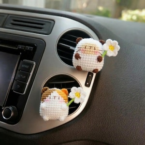 Pode incluir: Duas decorações de ventilação de carro de crochê feitas à mão. Uma é branca com detalhes marrons, a outra é marrom e laranja com detalhes brancos. Cada uma segura uma flor branca com centro amarelo.