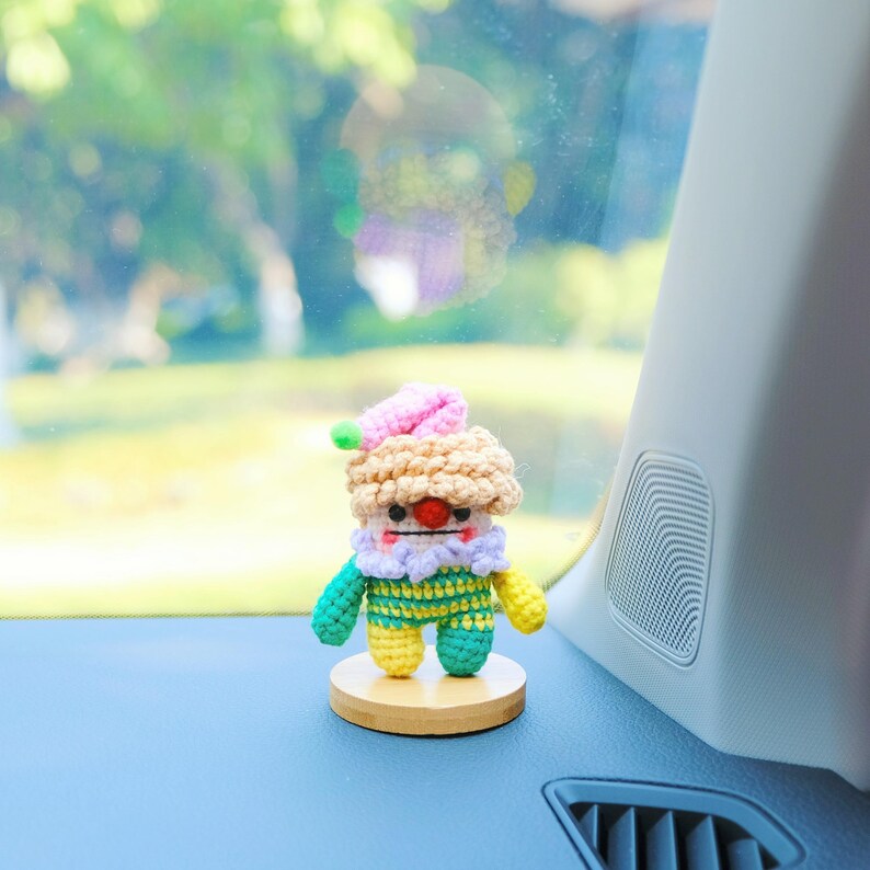 May include: A handmade crochet clown doll with a pink hat, beige hair, and a red nose. The doll has a colorful outfit with green, yellow, and blue accents. It is mounted on a small wooden base, perfect for display.