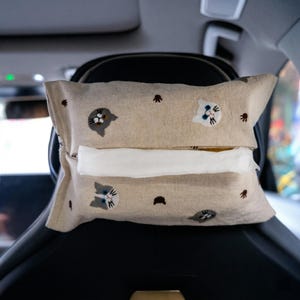 May include: Beige car tissue box cover with embroidered cat faces and small brown accents. The tissue box cover is rectangular and has a slot for tissue dispensing. The cover is placed on a car seat headrest.